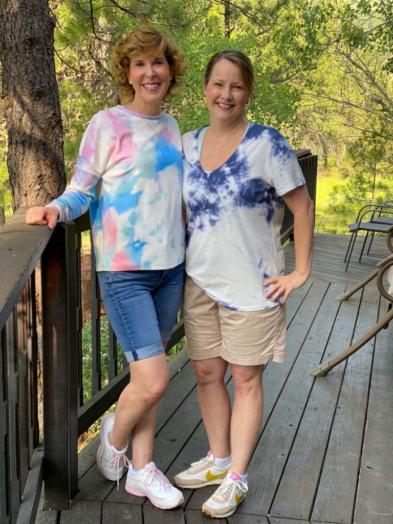 two women dressed in tie-dye shirts standing on the deck of a cabin in the woods