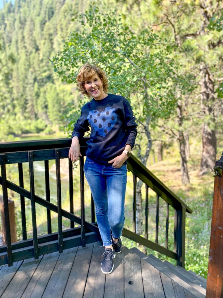 woman wearing black sweatshirt with blue heart standing on deck of a cabin in the woods