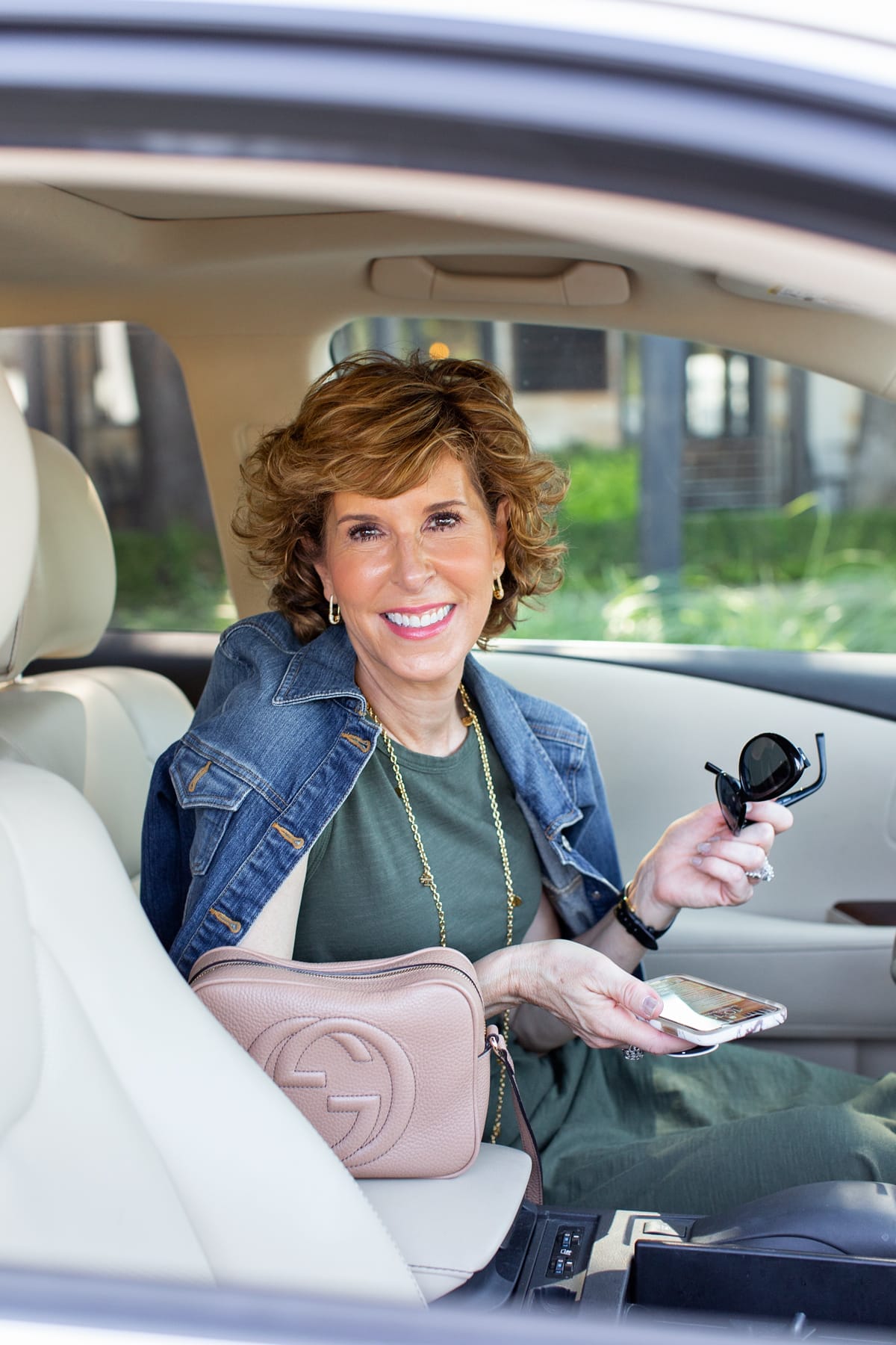 woman in green dress and denim jacket sitting in her car holding her phone looking at camera