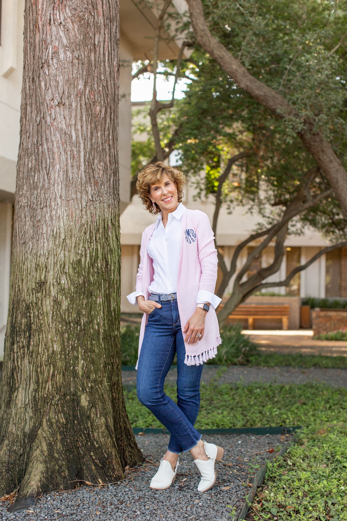 woman in pink cardigan with one hand in jeans pocket standing next to a tree trunk looking at the camera