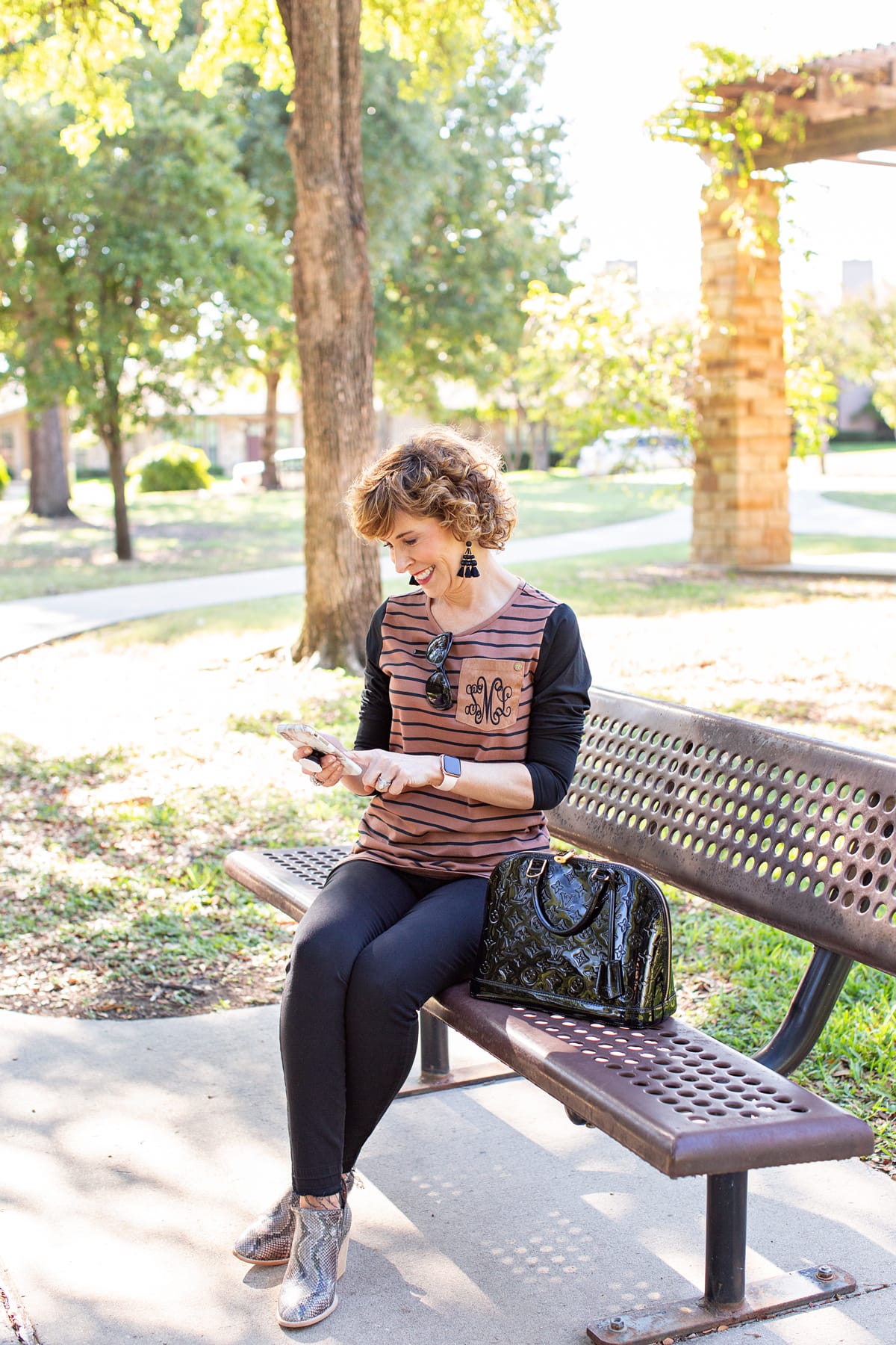 woman over 50 in brown and black outfit sitting on park bench looking at her phone