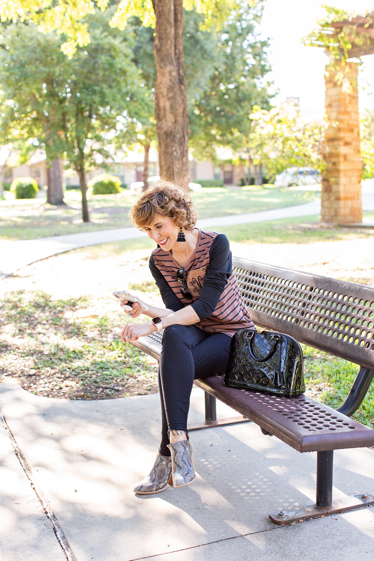 woman over 50 in brown and black outfit sitting on park bench looking at her phone