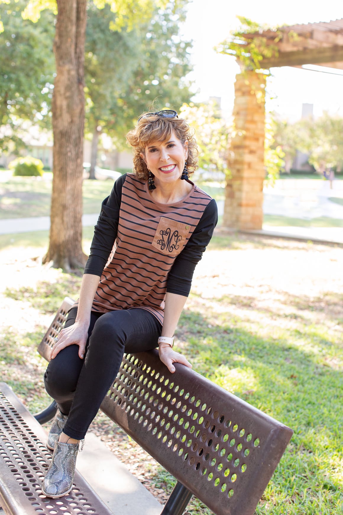 woman over 50 in brown and black outfit sitting on park bench looking at her phone