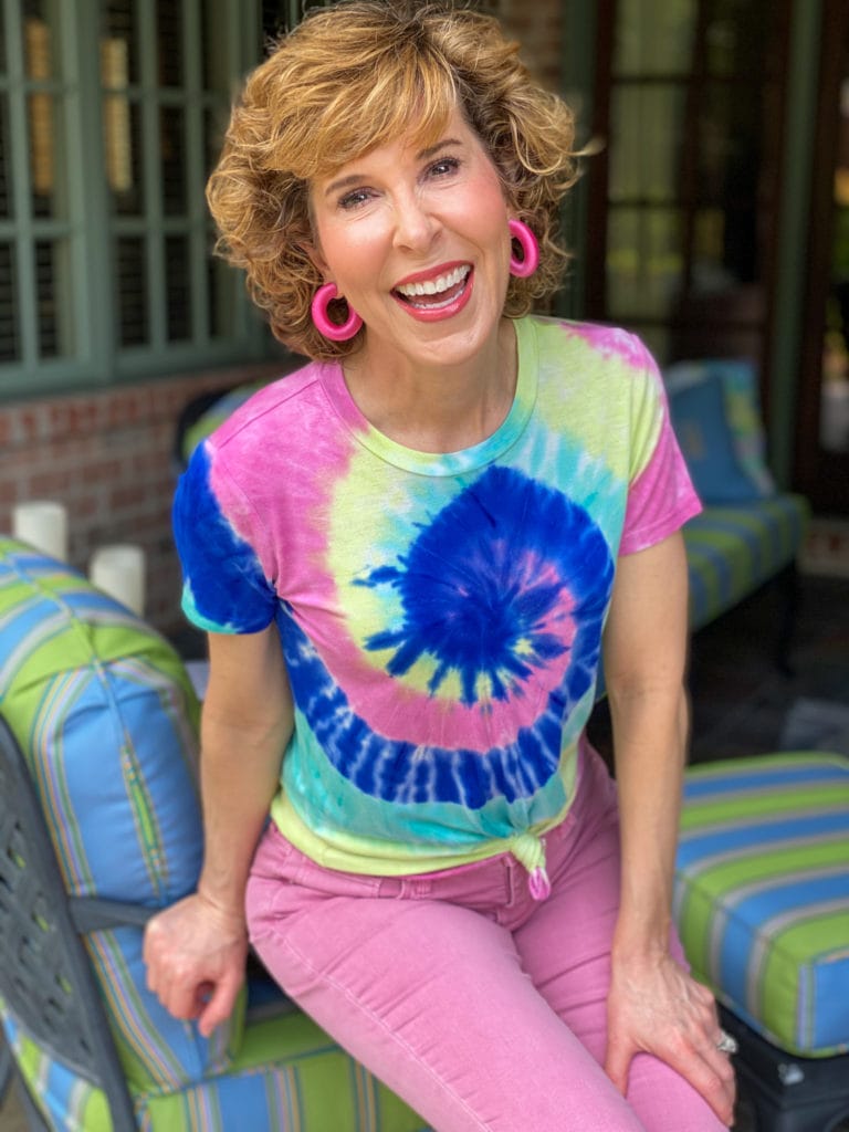 woman sitting on the edge of a chair wearing tie dye tee and bright pink earrings