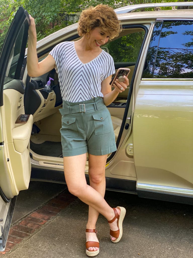 woman in green striped shirt and green shorts standing by car looking at cell phone