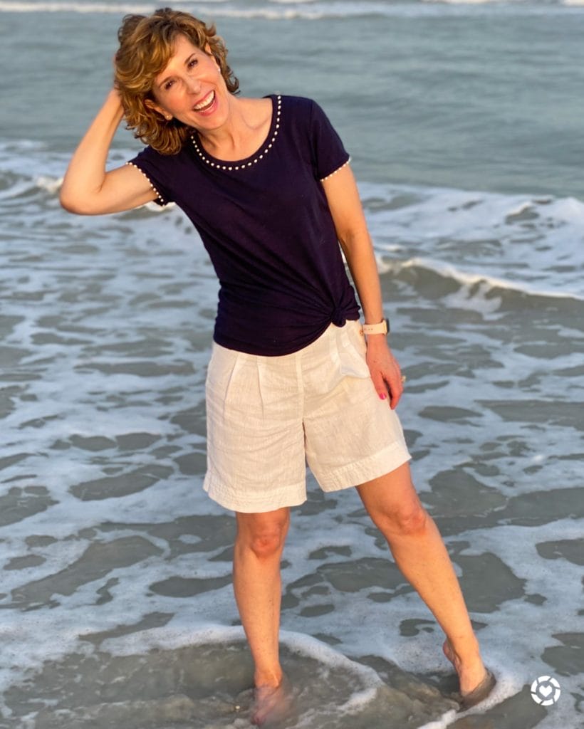 woman in navy tee and white linen shorts standing in the shallow water at the beach