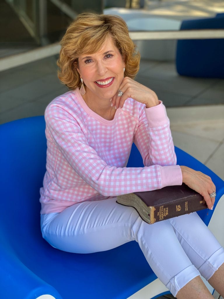 woman in pink gingham sweatshirt, white jeans, pink sneakers holding her Bible and sitting in a blue chair