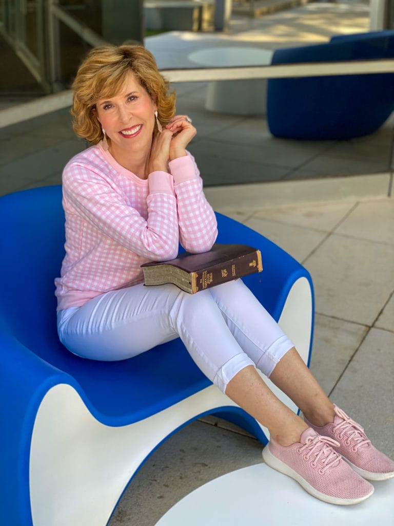 woman in pink gingham sweatshirt, white jeans, pink sneakers holding her Bible and sitting in a blue chair