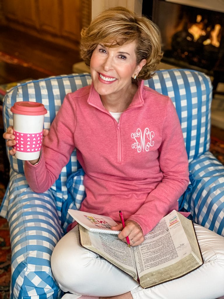 woman sitting in a blue and white gingham chair dressed in a pink monogrammed pullover and white jeans holding a pink and white coffee cup and with a bible and a journal in her lap praying for her adult children