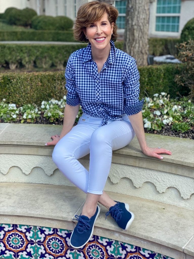 woman wearing talbots blue and white gingham shirt and white jeans with navy allbirds tree runner sneakers sitting on a wall