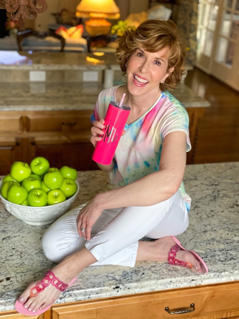 woman wearing tie dye shirt and white jeans with pink sandals holding a Marley Lilly pink tumbler sitting on a counter