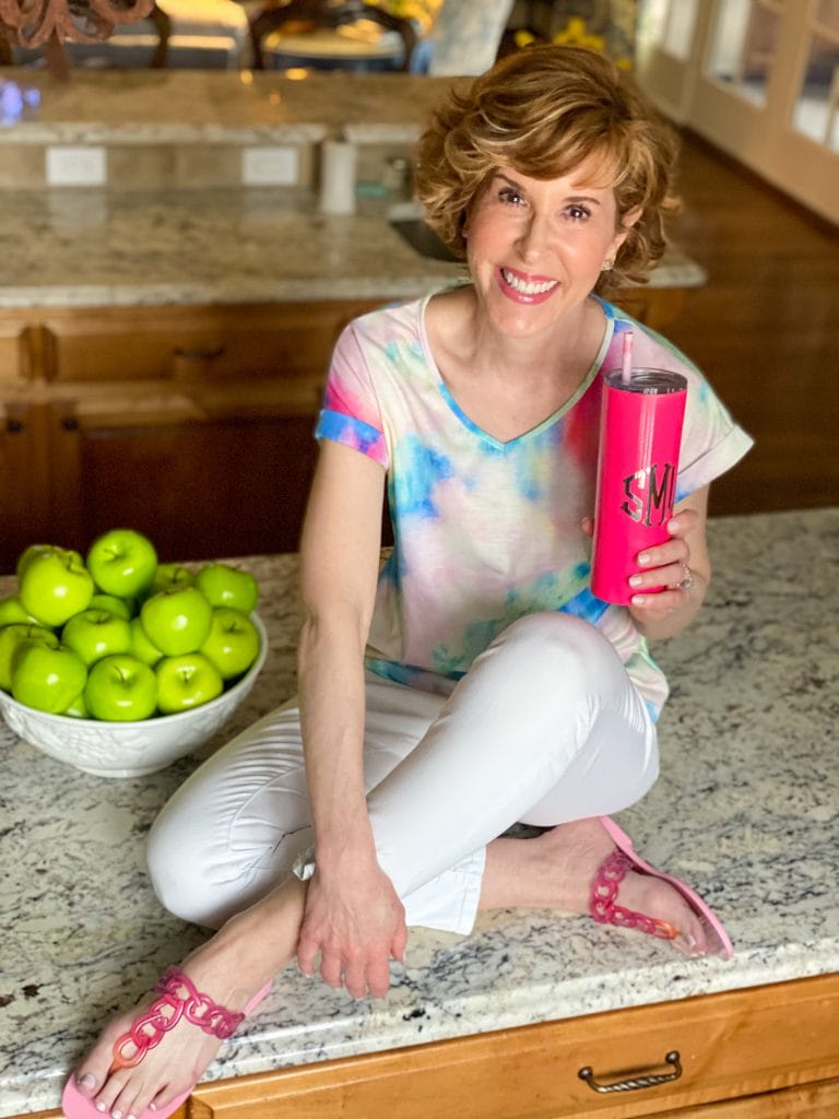 woman wearing tie dye shirt and white jeans with pink sandals holding a Marley Lilly pink tumbler sitting on a counter