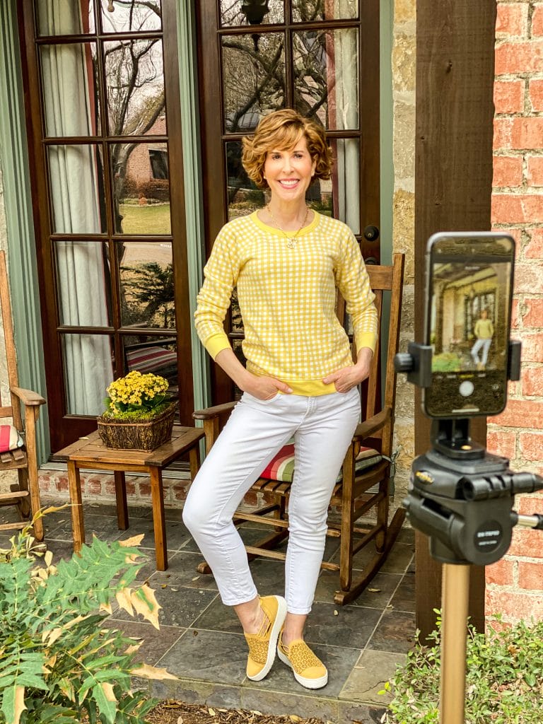 woman wearing draper james gingham and white sweatshirt and white jeans and yellow jambu sneakers standing on a front porch viewed through an iphone