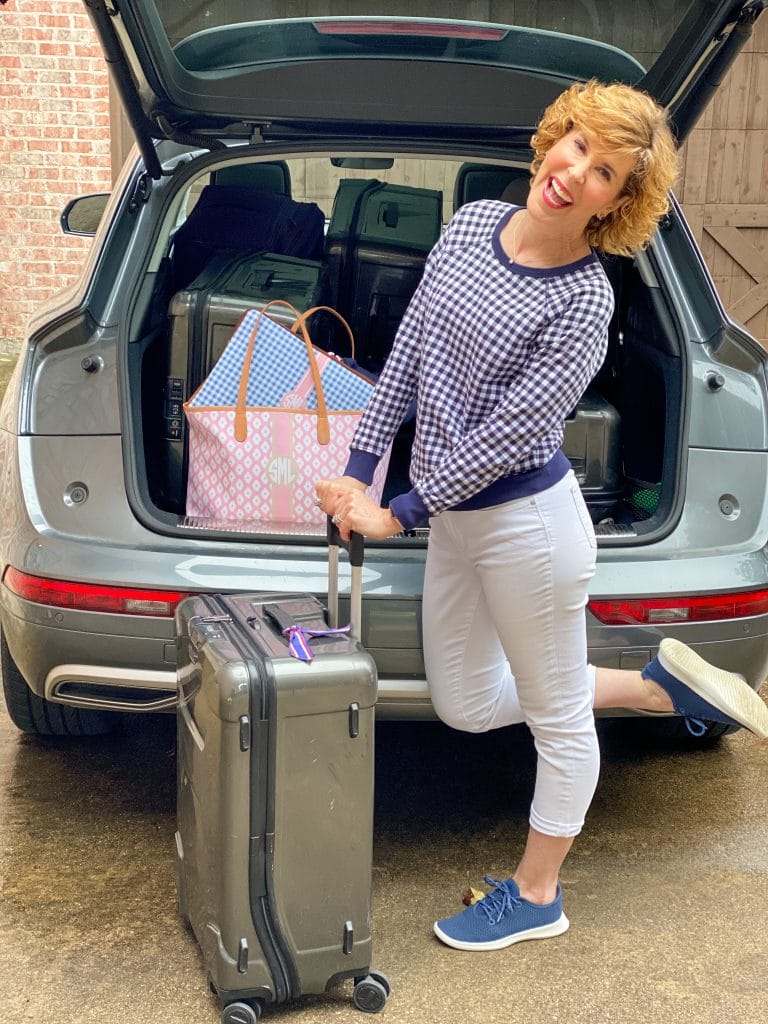 woman wearing draper james navy and white gingham sweatshirt and white crop jeans with navy allbirds tree runners standing with luggage behind a car with the trunk up