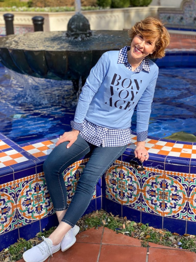 over 50 woman wearing Talbots blue bon voyage sweater over a blue and white gingham button down with jeans and sneakers sitting on the edge of a colorful fountain