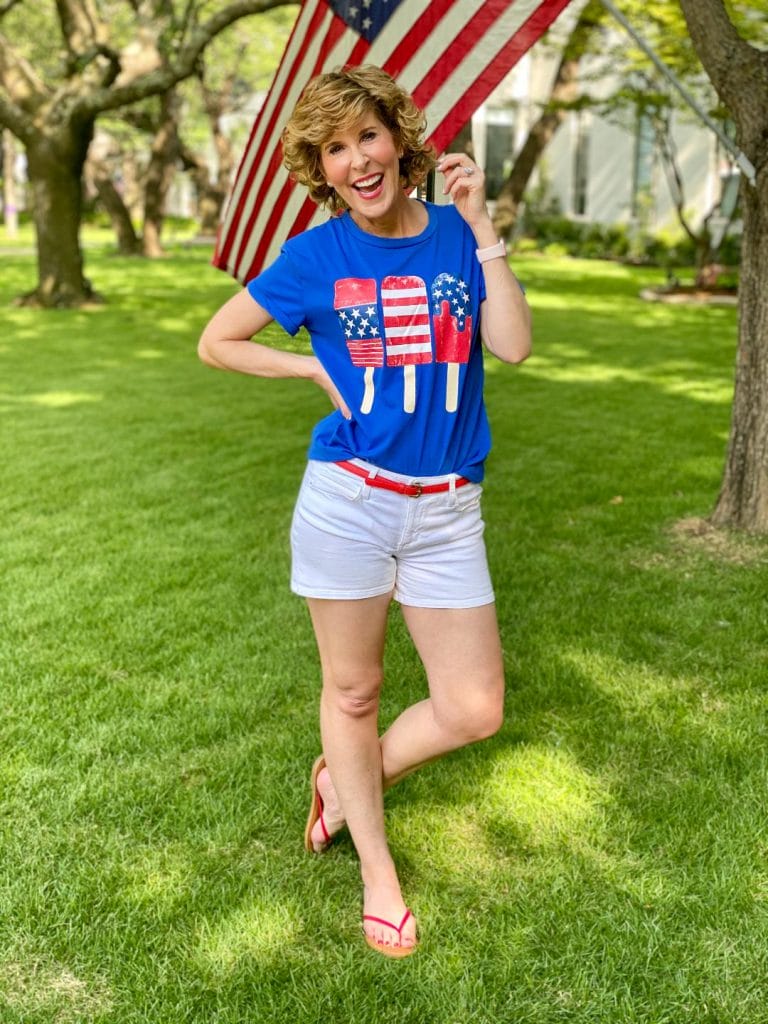 woman wearing Amazon patriotic blue tee with red white and blue popsicles and white shorts standing by an American flag