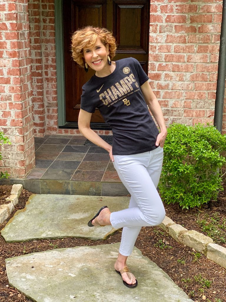 woman wearing black CHAMPS tee and white jeans standing on one foot in her front yard