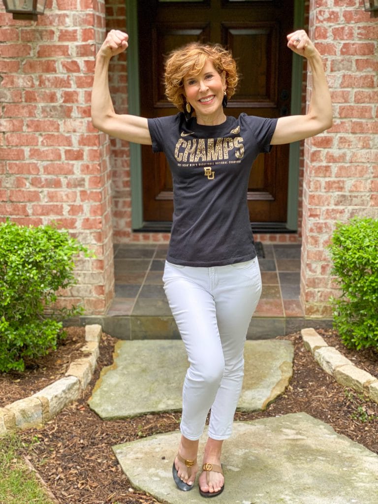 woman wearing black CHAMPS tee and white jeans standing in her front yard flexing her muscles