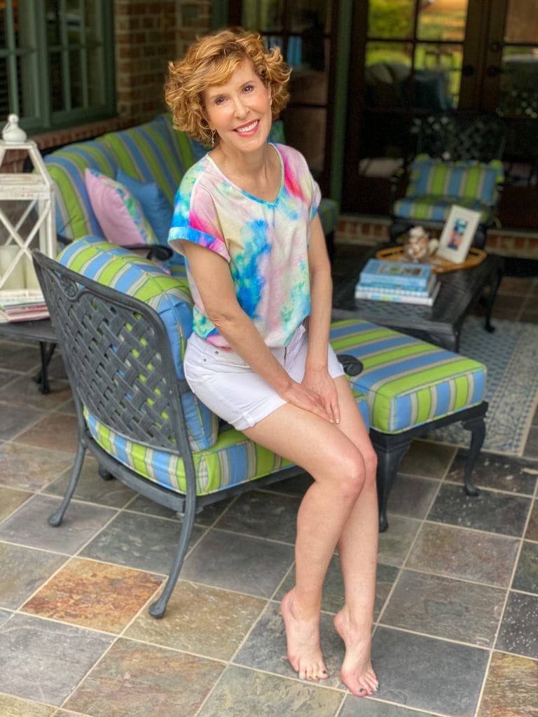woman over 50 wearing tie die tee and white shorts sitting on the arm of a chair with her back patio behind her