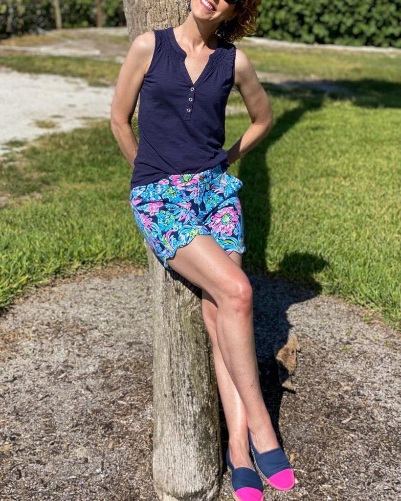 woman wearing jcrew factory navy blue rattan earrings, lilly pulitzer navy blue essie tank top and 7" darci knit shorts learning against a palm tree.