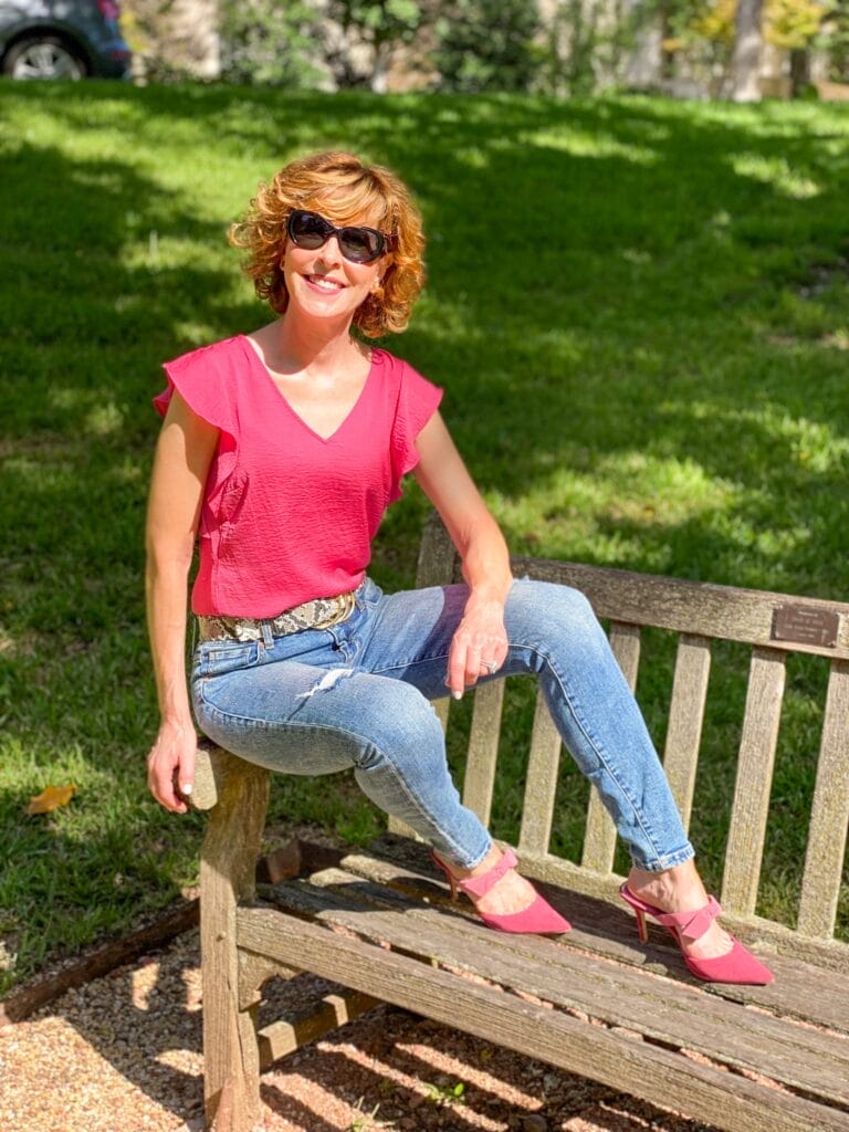 woman over 50 sitting on a park bench wearing cabi cinch skinny jeans, serpent belt, rose top, cheeky topper