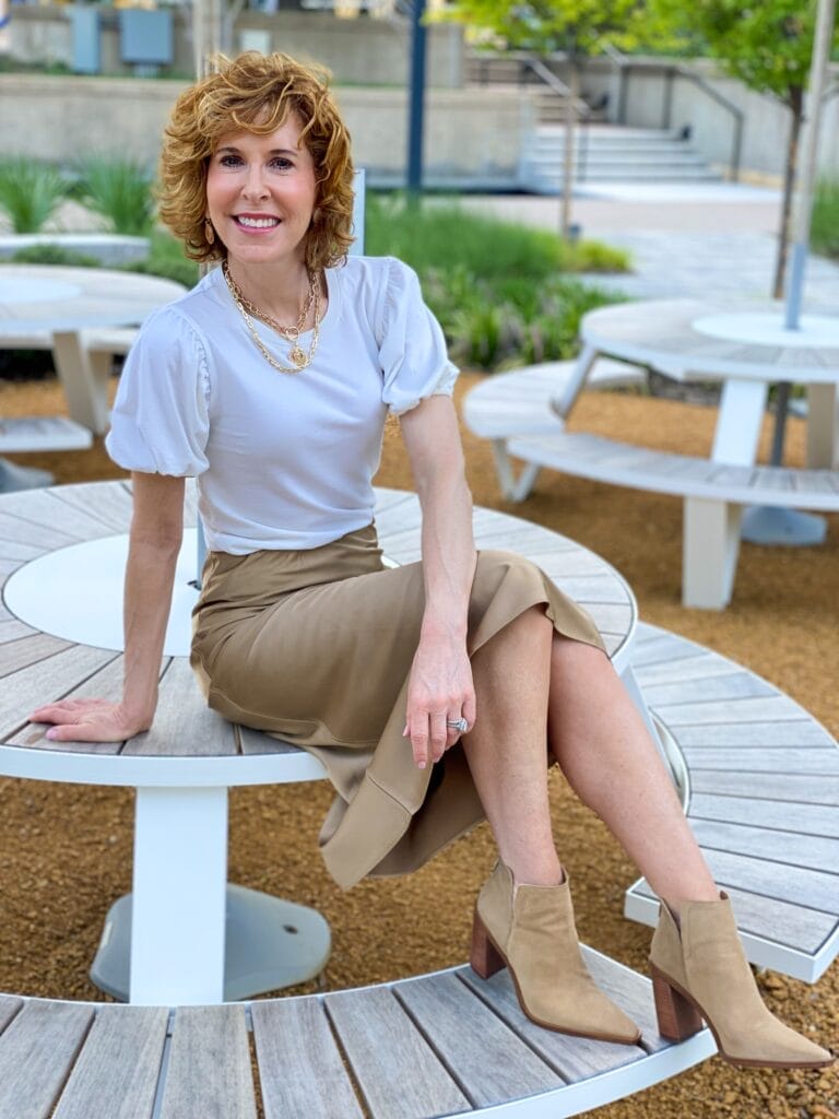 woman wearing puff sleeve tee with tan slip skirt and tan booties sitting on a table