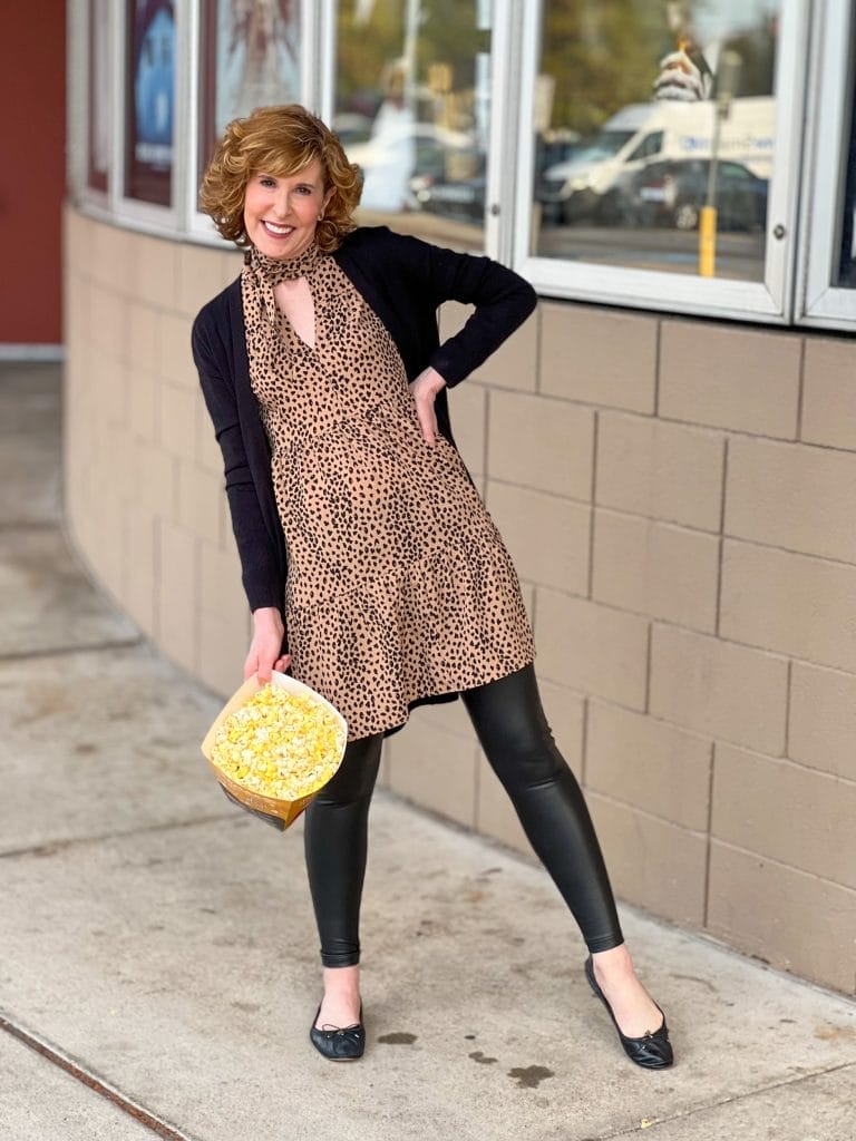 woman over 50 wearing nordstrom halogen Tie Neck Tiered Long Sleeve Dress, nordstrom brand faux leather leggings, nordstrom halogen open front pocket cardigan holding a popcorn bucket standing in front of movie posters