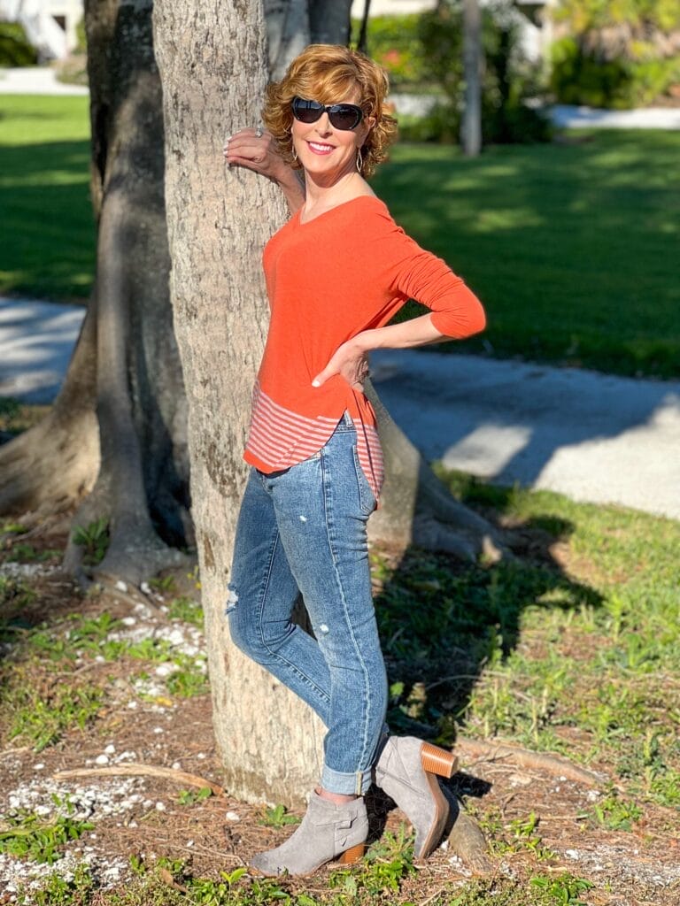 woman standing by a tree wearing cabi hangout tee and cinch skinny jeans with sunglasses and gray booties