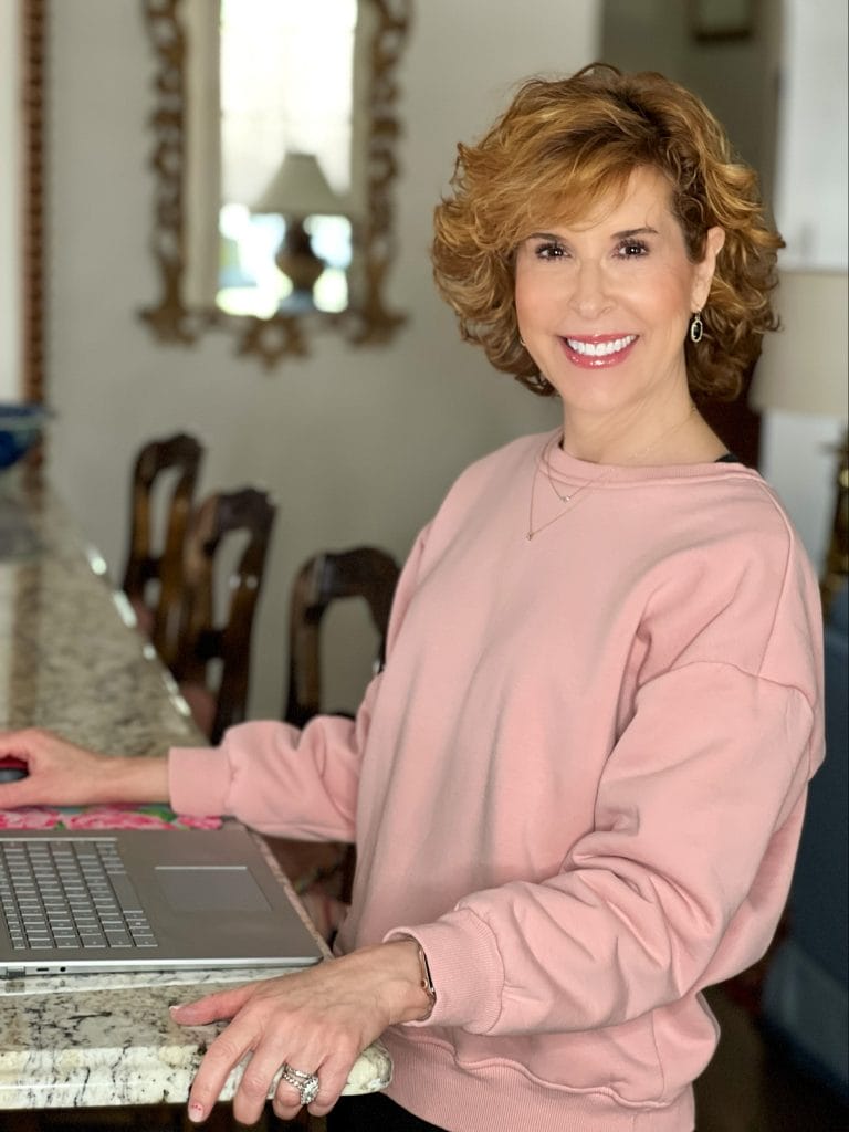 woman over 50 wearing amazon pink fleece-lined sweatshirt and black joggers standing up working on a laptop
