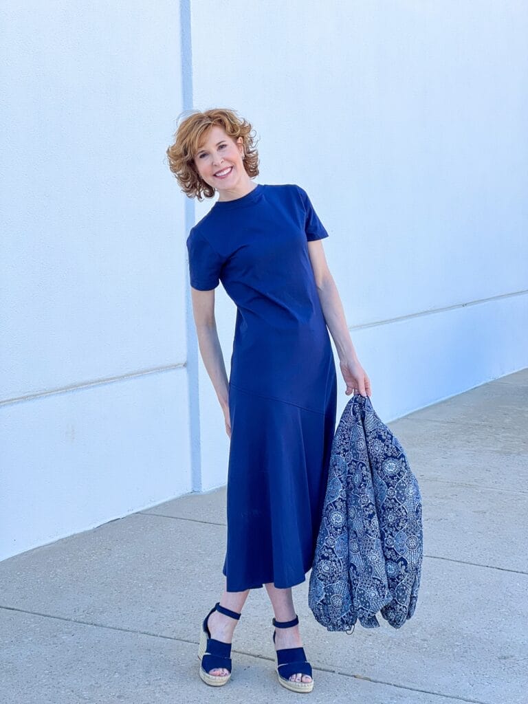 woman over 50 standing against white wall wearing Caslon® A-Line T-Shirt Cotton Maxi Dress from Nordstrom with navy wedges and holding a navy quilted patterned jacket