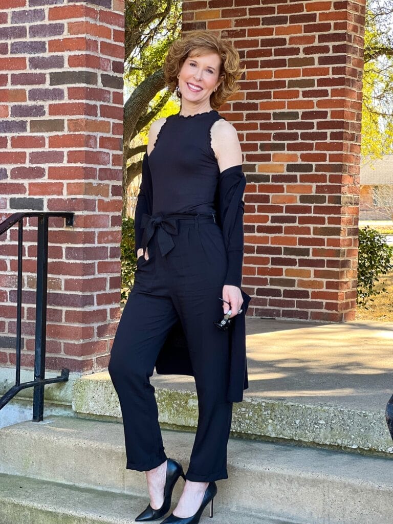 woman over 50 posing on staircase wearing black Treasure & Bond Paperbag Waist Pants, wearing Ruffle Rib Tank and an open front cardigan over her arms as she transitions her wardrobe from spring to winter