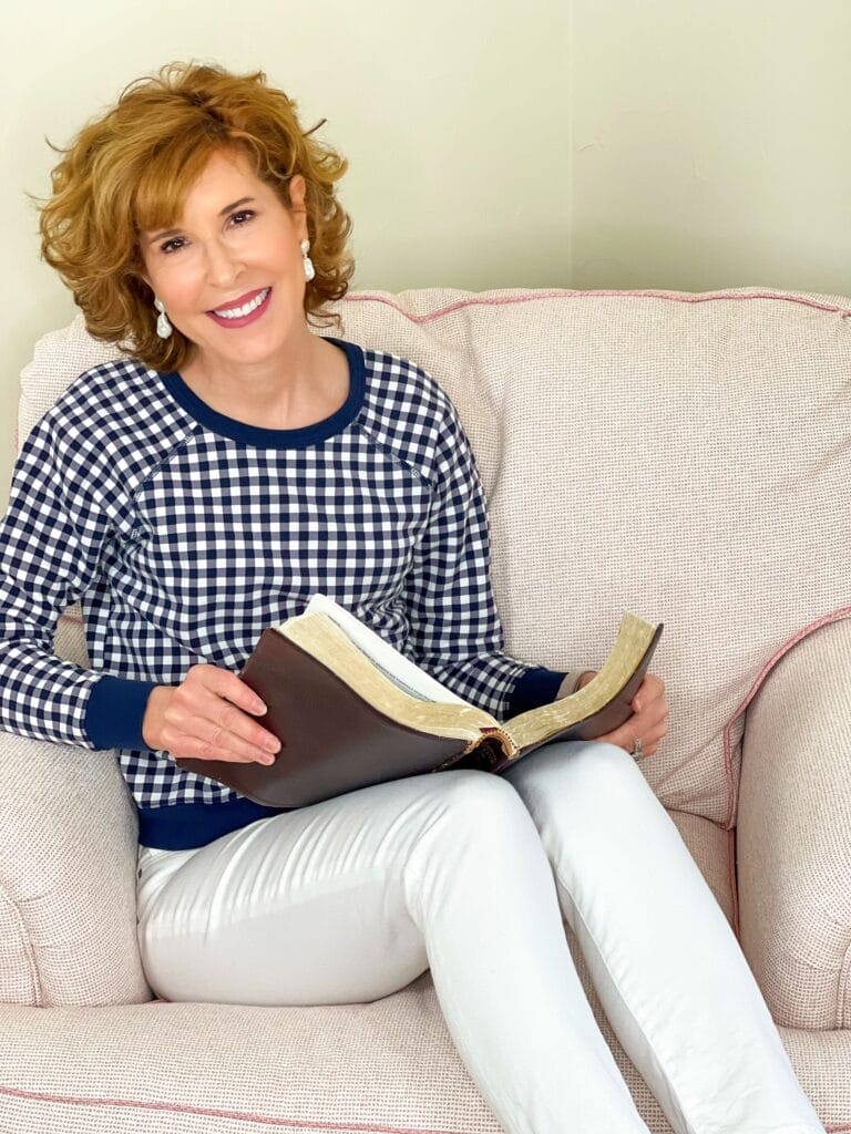 woman sitting in pale pink chair wearing Draper James navy and white gingham pullover and white jeans with a Bible in her lap