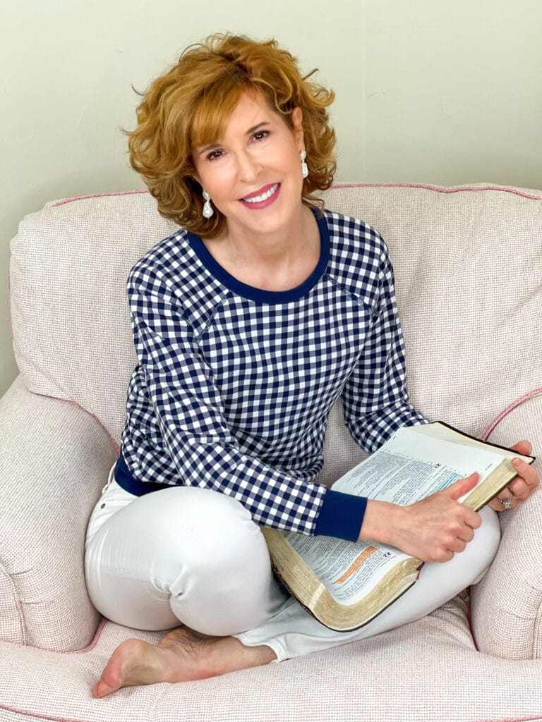 woman sitting in pale pink chair wearing Draper James navy and white gingham pullover and white jeans with a Bible in her lap praying for adult children questioning their faith