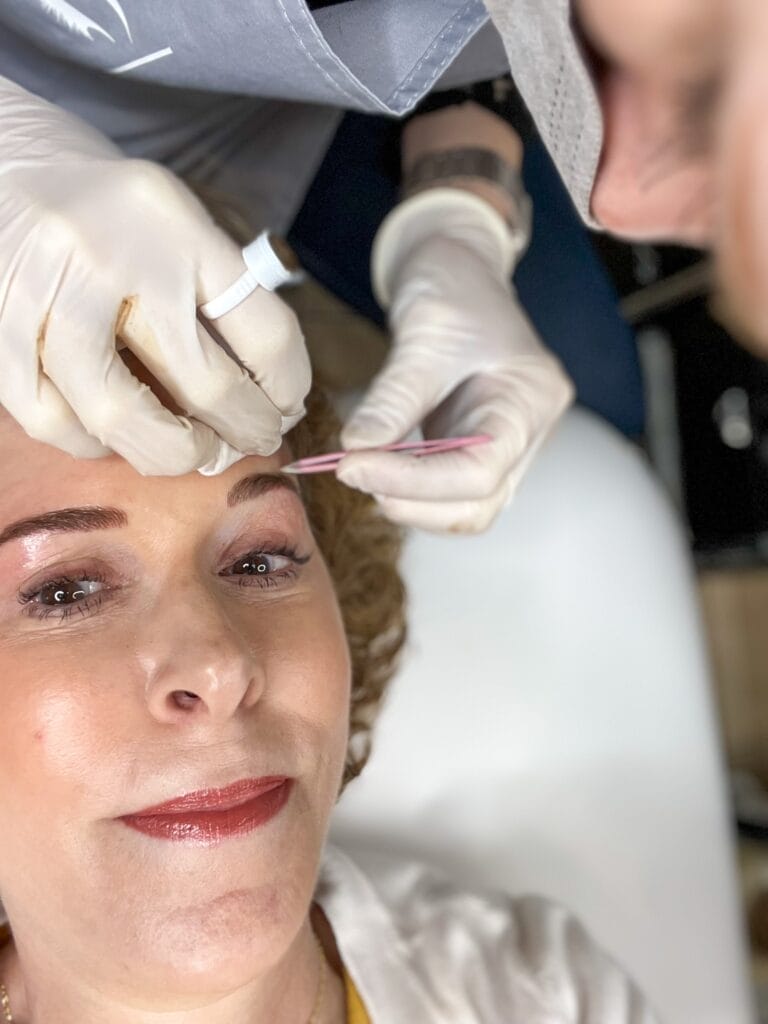 woman having her brows tweezed after microblading by pink tweezers and two gloved hands 