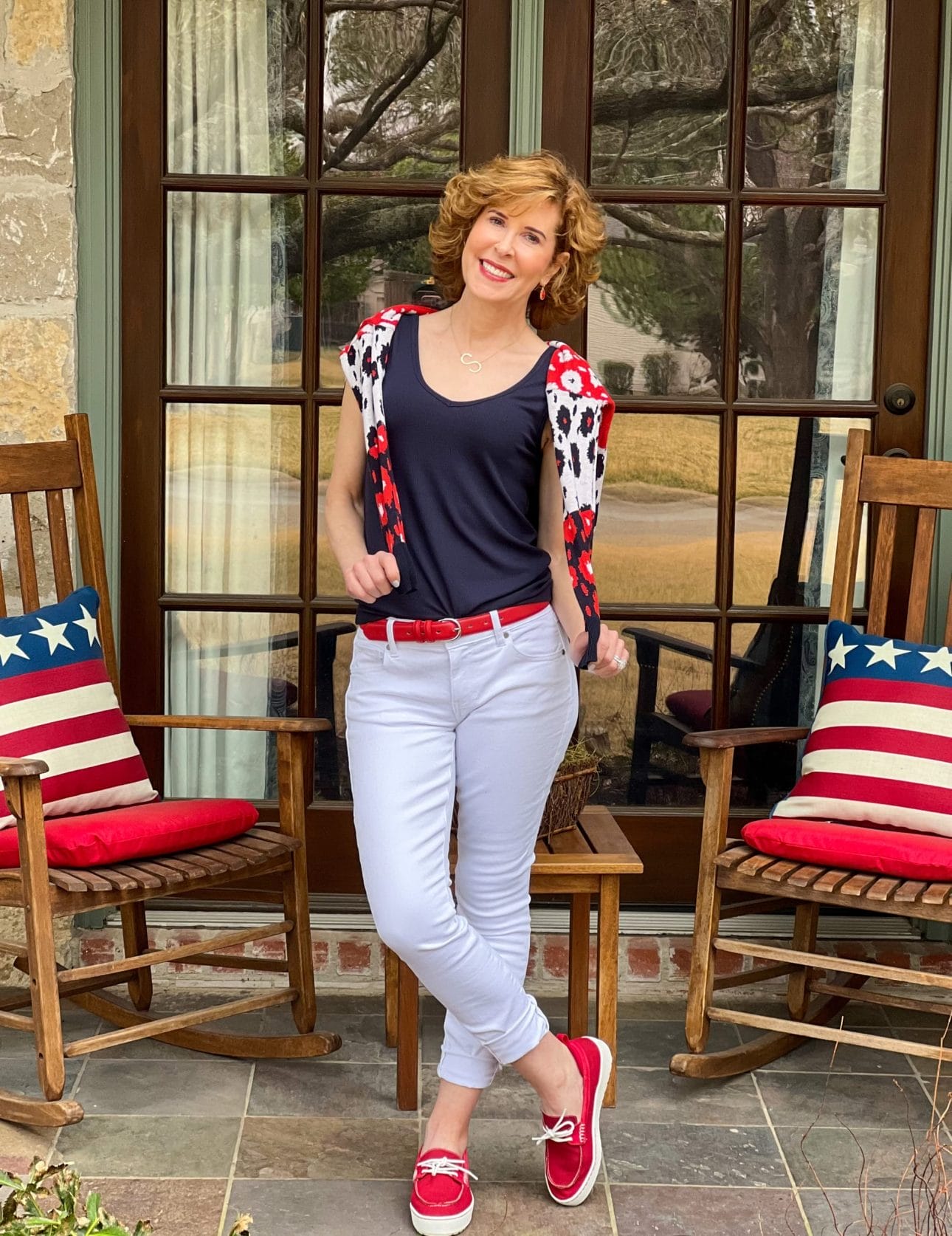 woman over 50 standing on a front porch with rocking chairs and patriotic pillows wearing from the 2022 cabi spring collection including white the skinny jeans, navy blue busy tank, and the upbeat cardigan on her shoulders with sleeves hanging down in front of her chest
