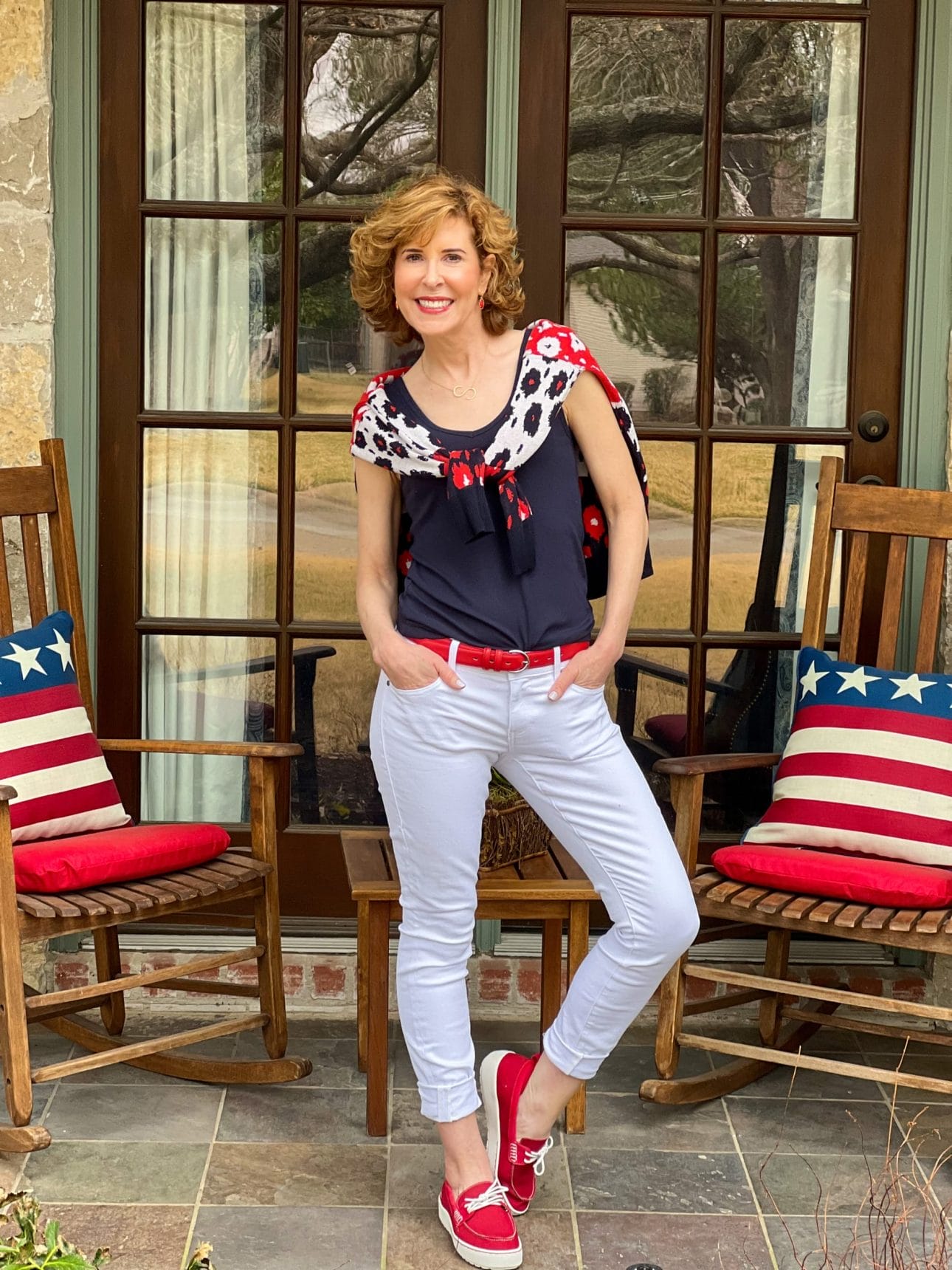 woman over 50 standing on a front porch with rocking chairs and patriotic pillows wearing from the 2022 cabi spring collection including white the skinny jeans, navy blue busy tank, and the upbeat cardigan tied around her shoulders