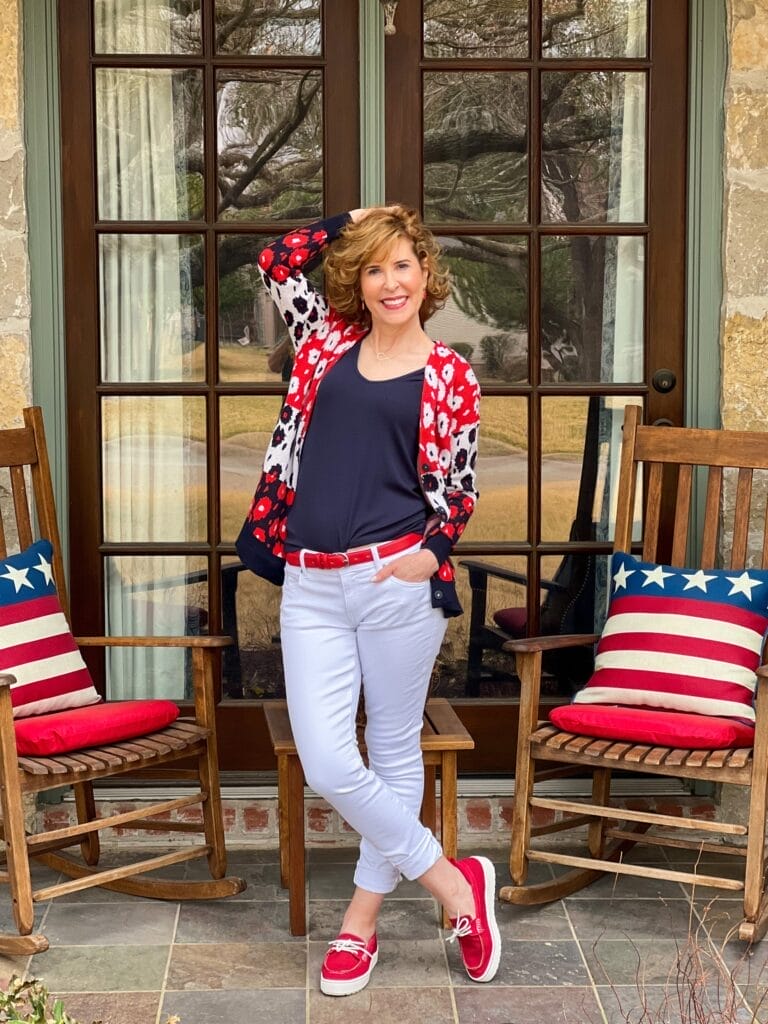 woman over 50 standing on a front porch with rocking chairs and patriotic pillows wearing from the 2022 cabi spring collection including white the skinny jeans, navy blue busy tank, and the upbeat cardigan