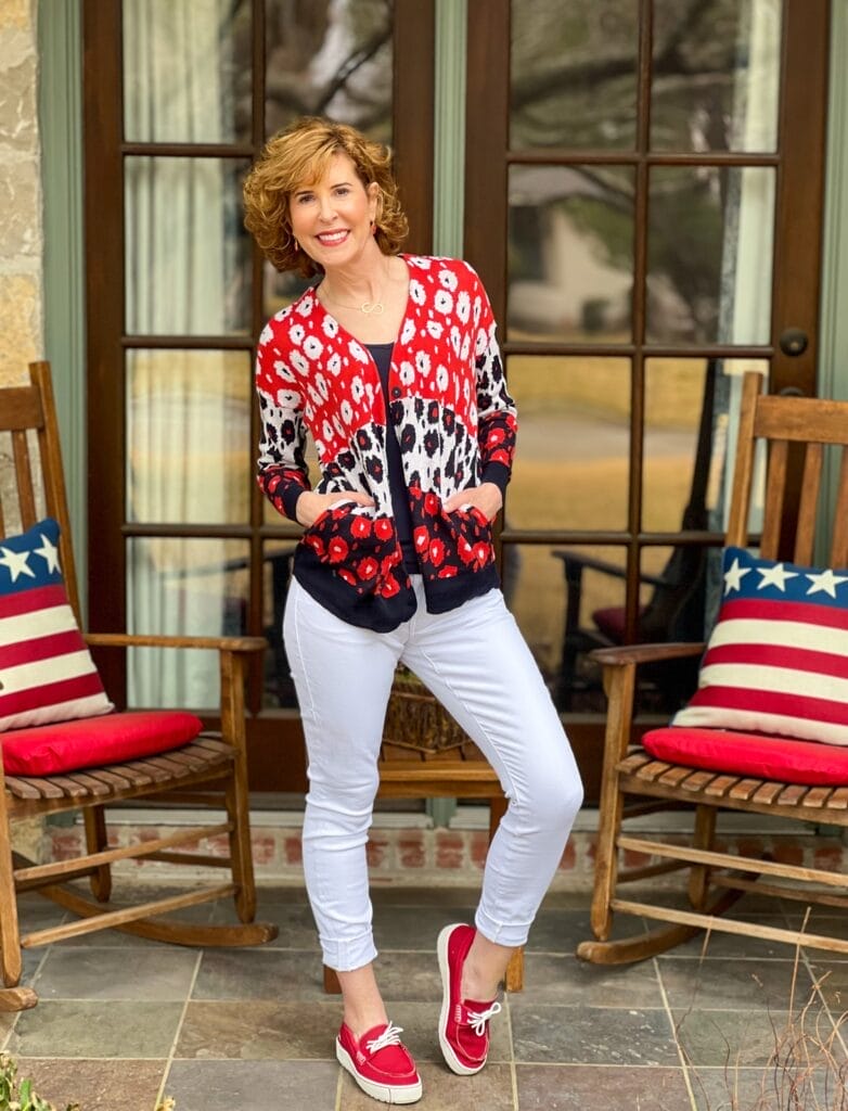 woman over 50 standing on a front porch with rocking chairs and patriotic pillows wearing from the 2022 cabi spring collection including white the skinny jeans, navy blue busy tank, and the upbeat cardigan on top with hands in the pockets