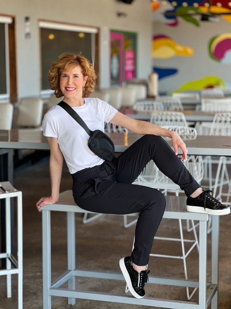 woman over 50 posing on a stool on a restaurant patio wearing black pants and a white tee with a black lululemon belt bag over her chest
