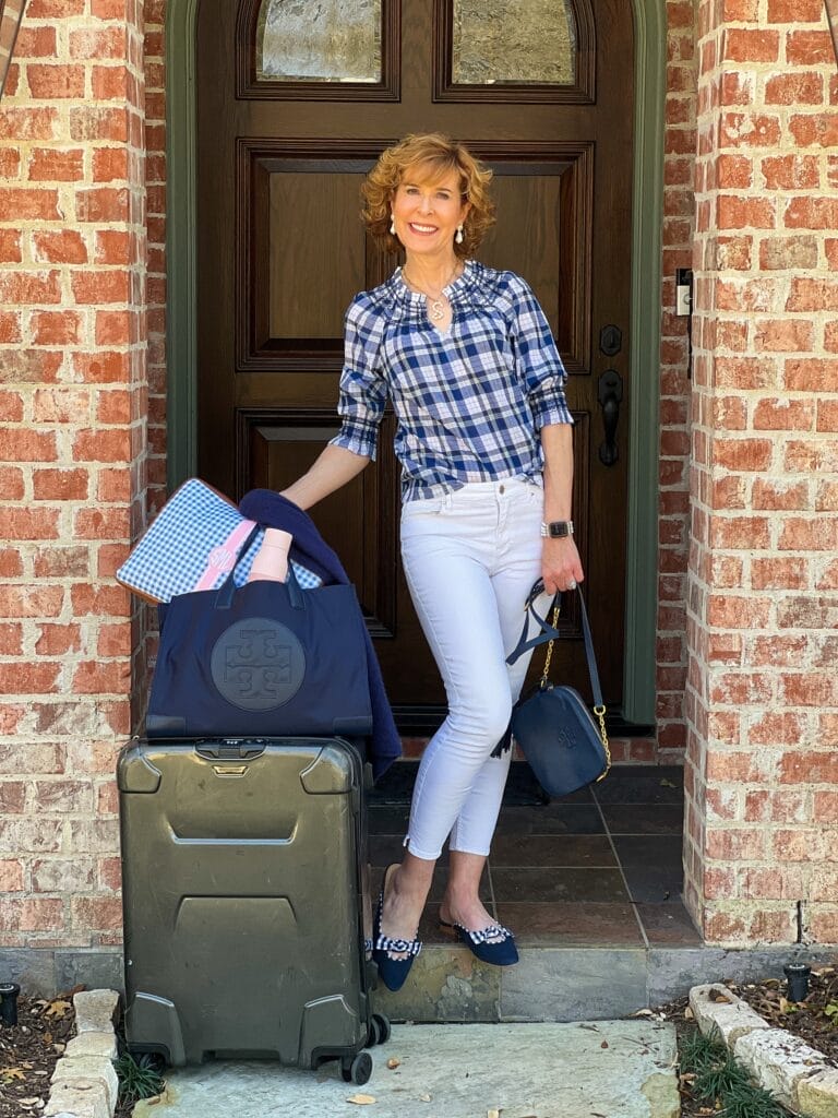 woman over 50 getting ready to leave on a trip wearing Talbots Plaid Smocked Blouse, chico’s no stain white jeans, and talbots Edison Gingham Suede Bow Mules standing alongside luggage