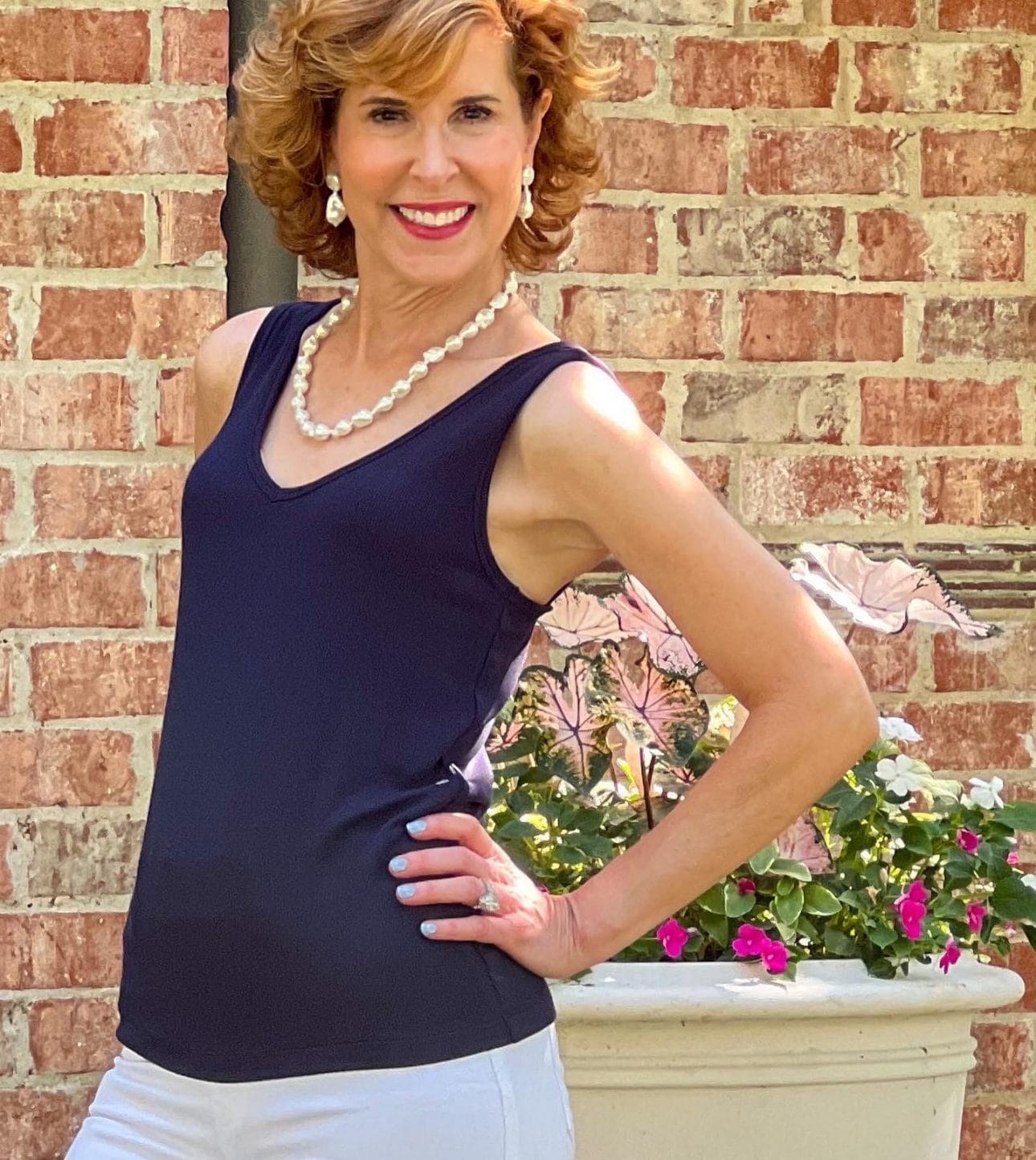woman over 50 standing with hand on her hipin front of a home wearing cabi spring style busy tank top with pearl jewelry