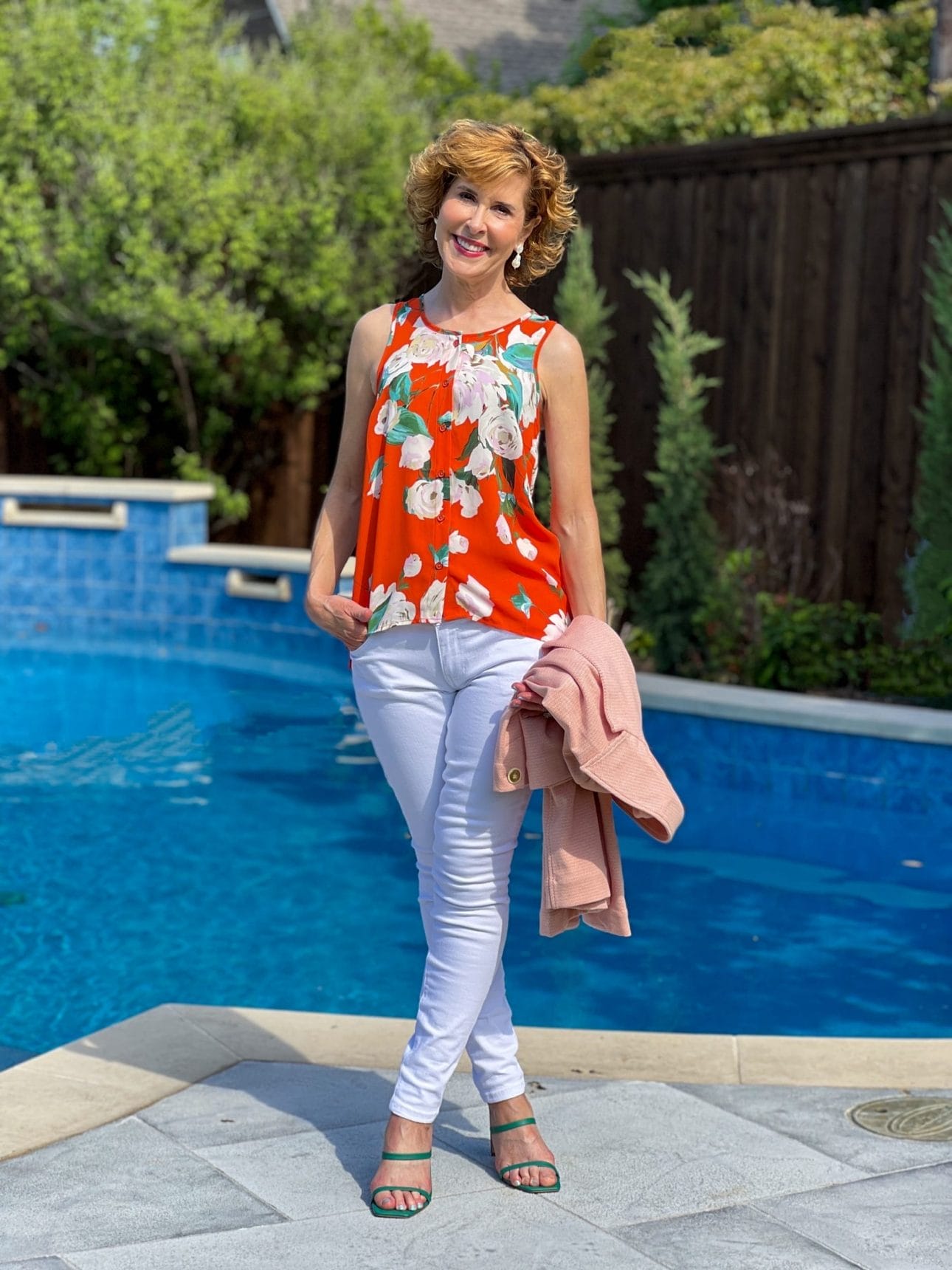woman posing in orange floral top and white skinny jeans by pool with her legs crossed