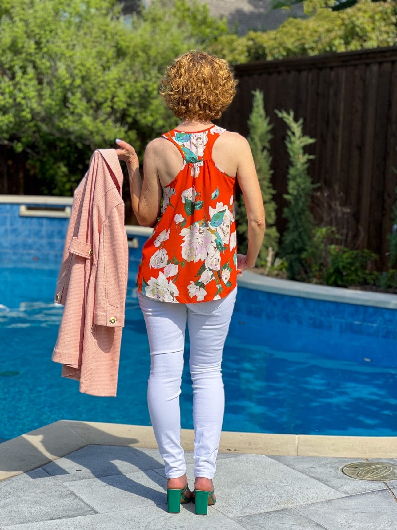 woman posing in cabi orange floral top and white skinny jeans by pool with jacket in her hand facing backwards