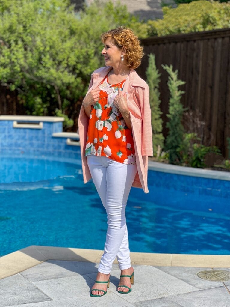 woman posing in orange floral top and white skinny jeans by pool with jacket over her shoulders looking sideways