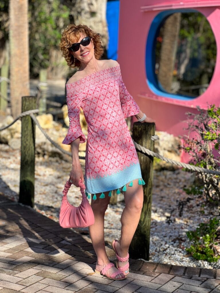 woman over 50 wearing a coral colored off the shoulder dress standing in front of coral building