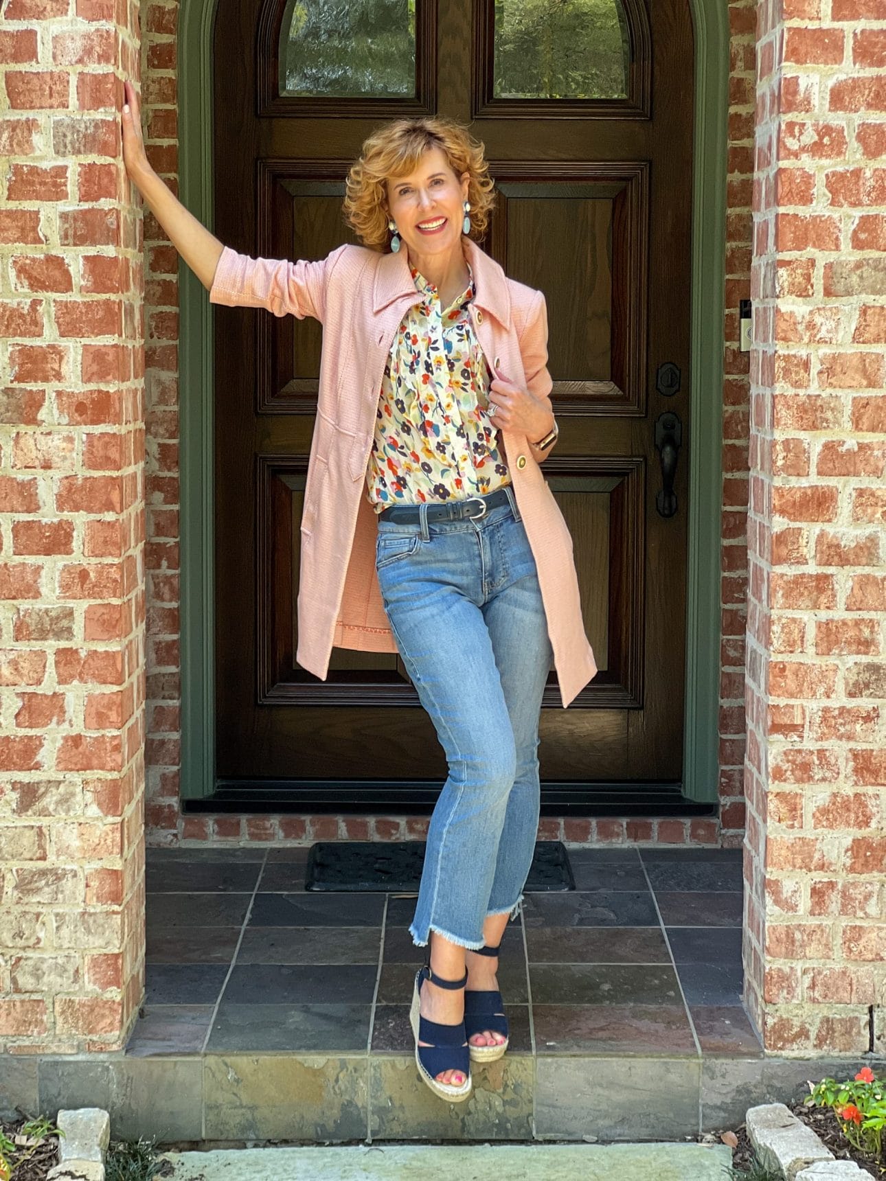 woman over 50 standing on porch wearing pink cabi take 5 topper over tux ruffle top and high low crop jeans