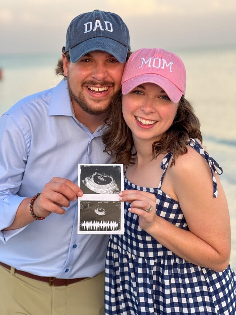 couple wearing mom and dad hats holding a sonogram picture and standing on the beach