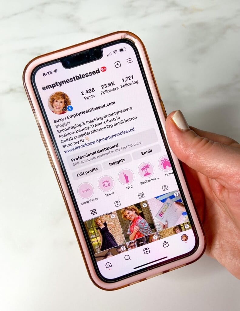 woman holding a phone showing the empty nest blessed instagram profile page on an iphone with a white marble background