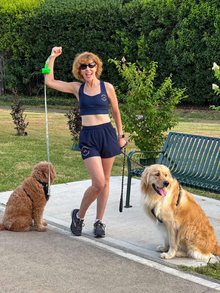 woman standing near a bench on a walking trail with 2 dogs on leash wearing mighty health sports bra & shorts making a muscle