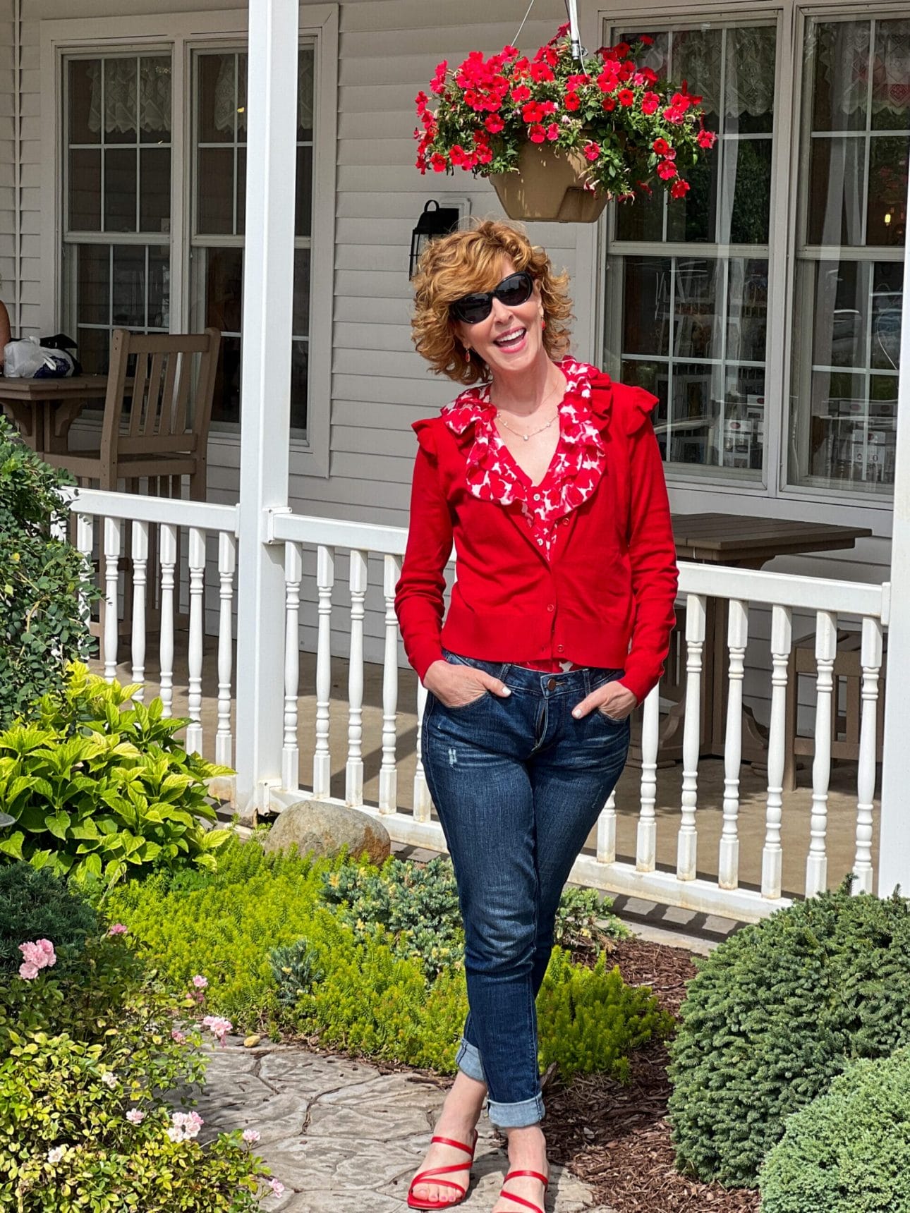 woman standing in front of a white porch railing wearing the rosy top, cavalier cardigan and button fly straight jeans from the cabi fall collection